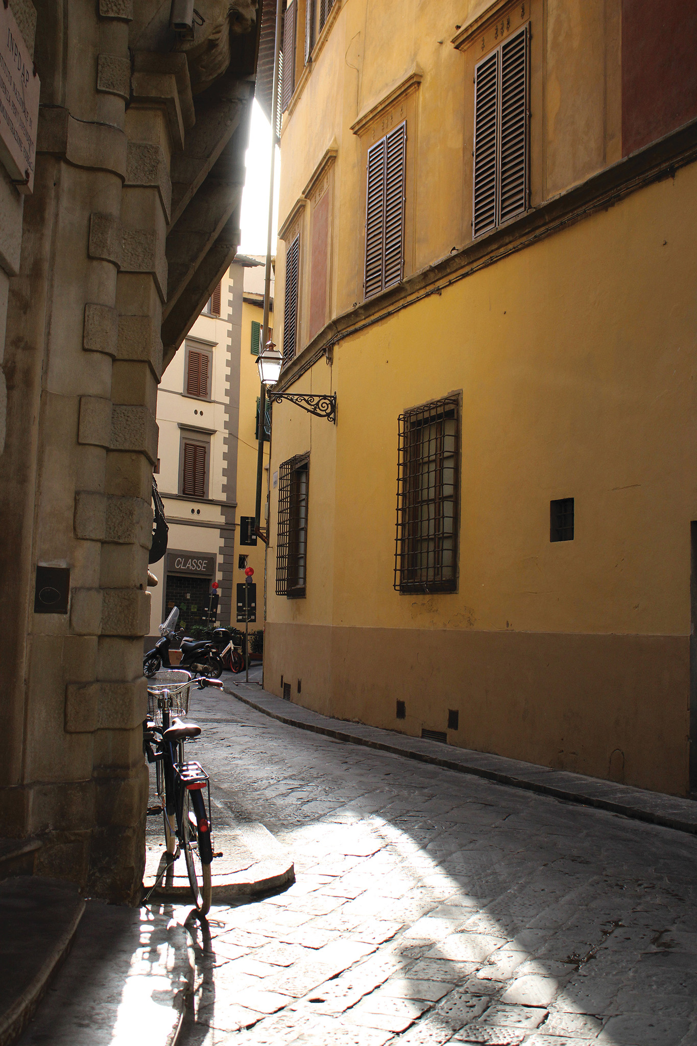 This acts as an example of an image before proper editing. It depicts a small alley with the first rays of sun shining through and illuminating a bike in the corner.