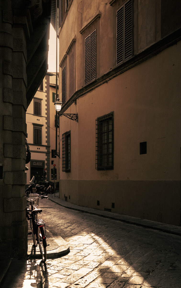 Image for photography page. It shows a dark alley with light filtering in and shining on a lone bike, bathing the alley in warmth.