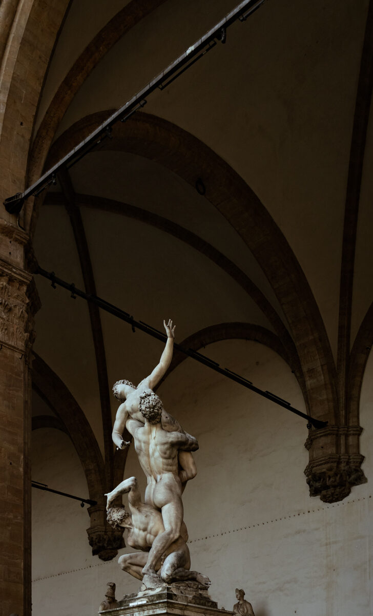 Image for photography page. A white marble statue of two men and a women reaching above each other sits underneath the arched roof of an old stone building.