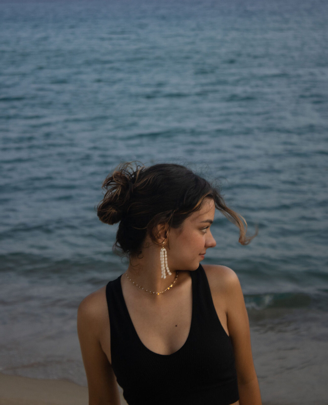 A teen girl in black top and pearl earrings stands at the edge of the water at a sandy beach as window blows through her hair.