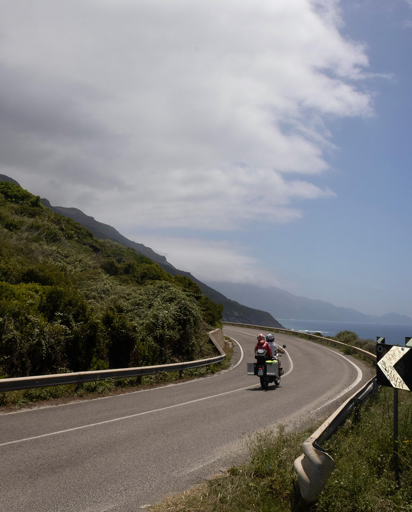 A motorcycle with two passengers drives down a winding road around the edge of a mountain.