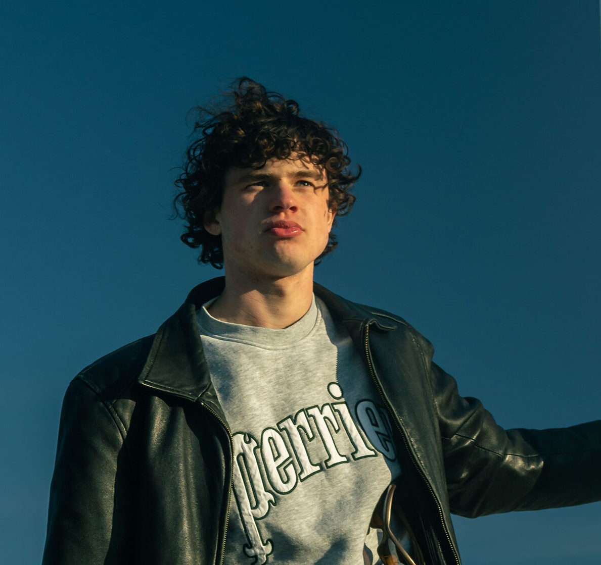 A teen boy in a leather jacket stares into the distance and the light from the setting sun hits one side of his face.