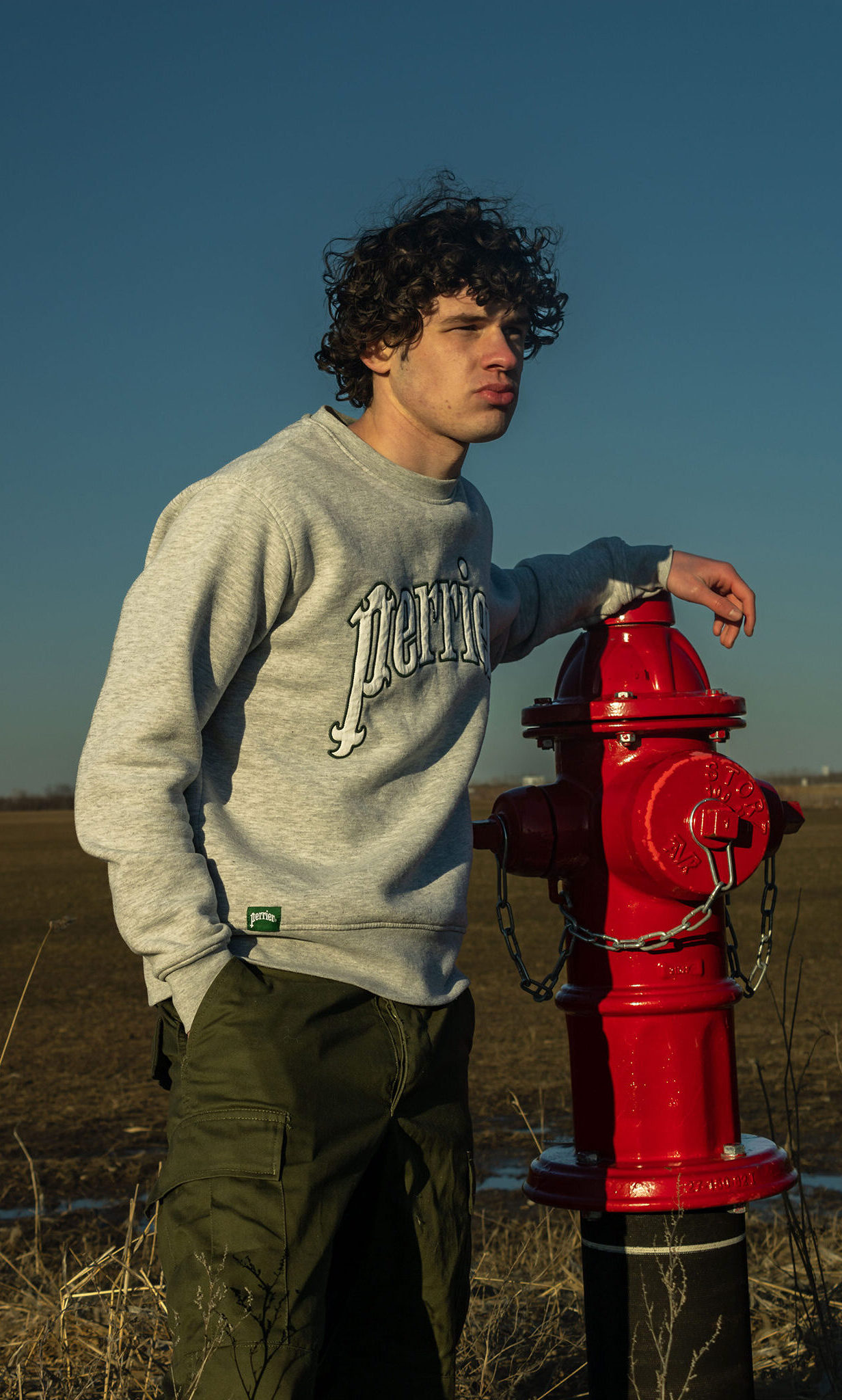 A teen boy in a grey sweatshirt stands in a field leaning against a bright red fire hydrant.