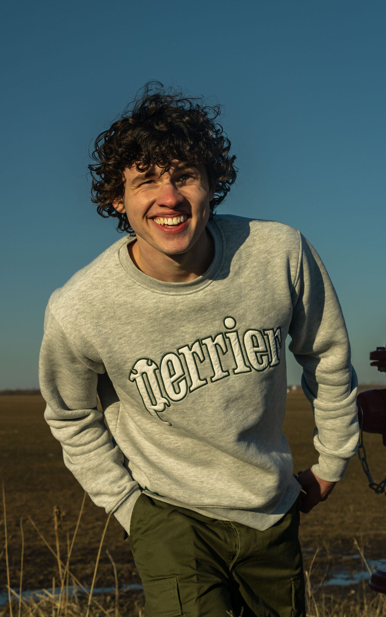 A teen boy in a grey sweatshirt stands in a field leaning towards the camera.