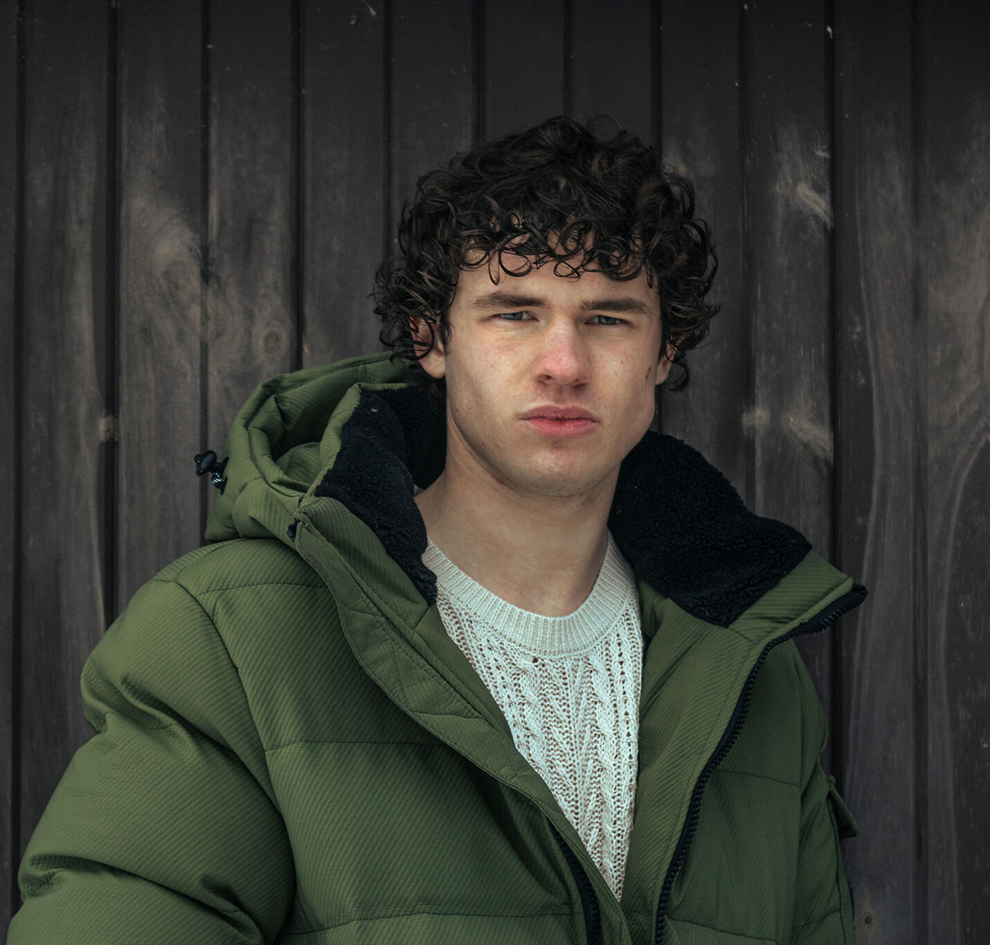 A teen boy with curly hair in a thick green winter coat and a white cable knit sweater stands in front of a barn door. 