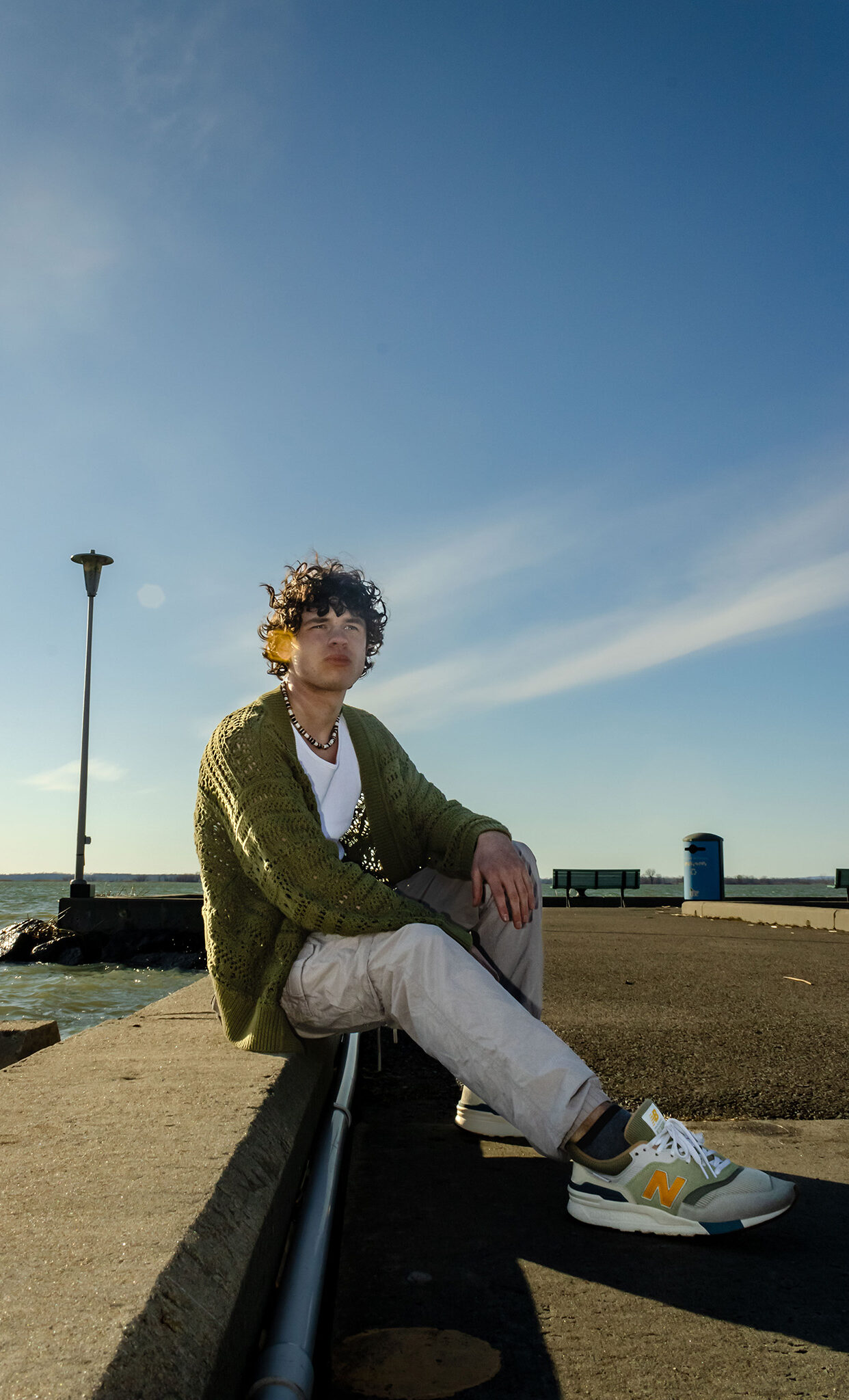 A teen boy with curl hair in a green cardigan sits at a peer on a sunny day.