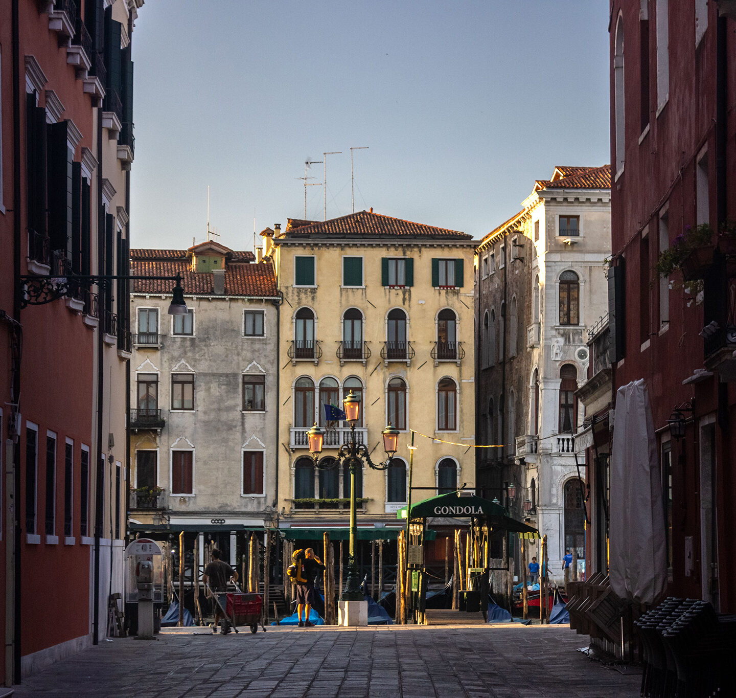 Street in Venice opens up to the canals where gondolas are lined up in the earl morning. The street is shrouded in darkness white the buildings on the opposite end of the canal gleam with light.
