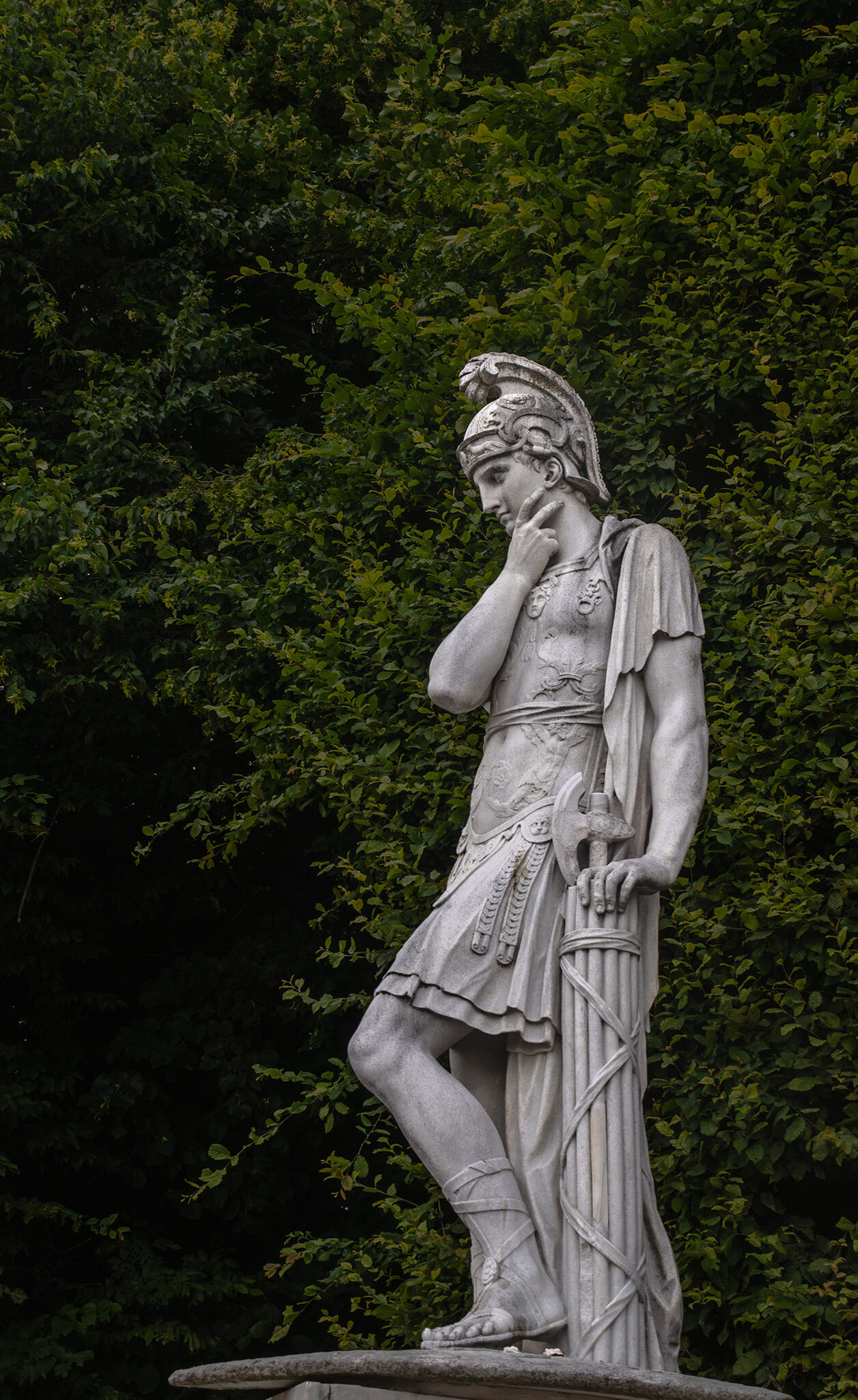 A grey statue stands pondering on a backdrop of greenery.
